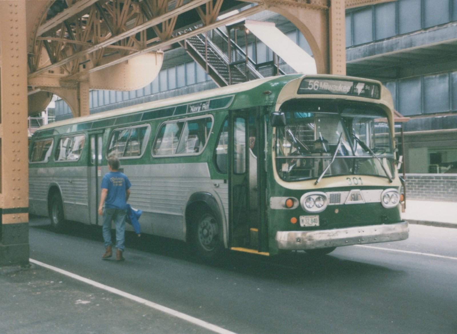 A front three-quarter view of a green and silver CTA bus numbered 301 on route 56 Milwaukee, parked under a metal elevated structure with a person walking beside it.