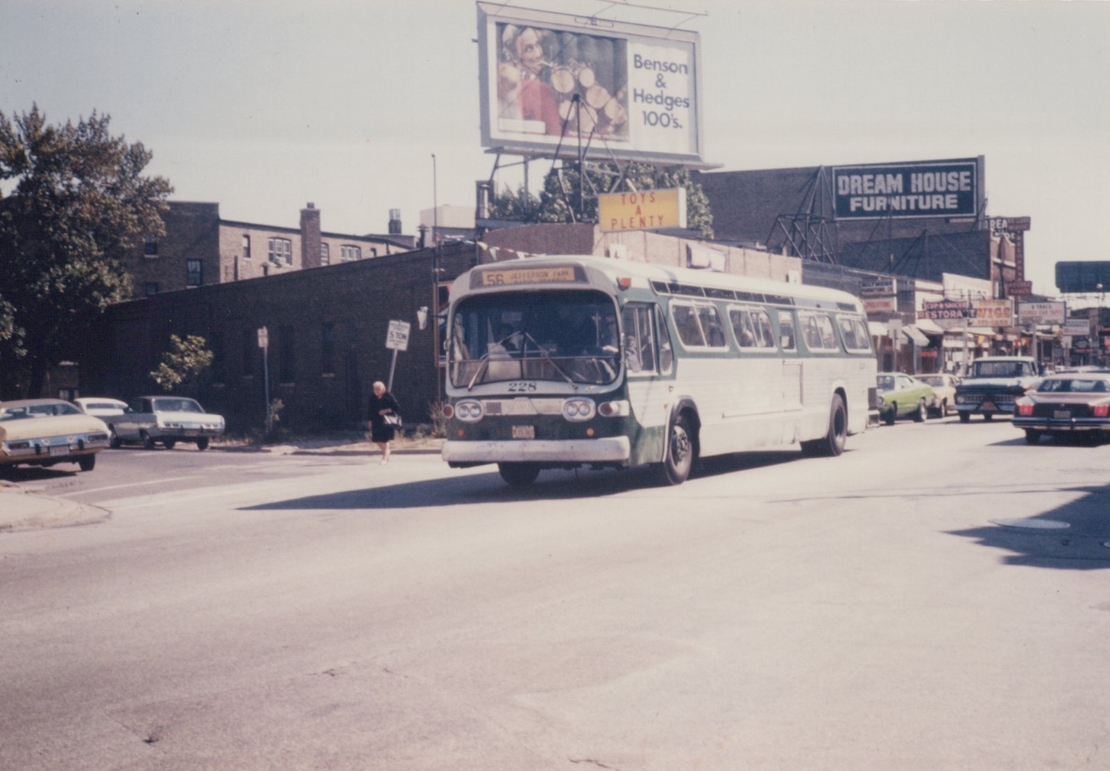 A vintage green and white city bus, number 228, turns a corner on a sunny street lined with 1970s cars and billboards.