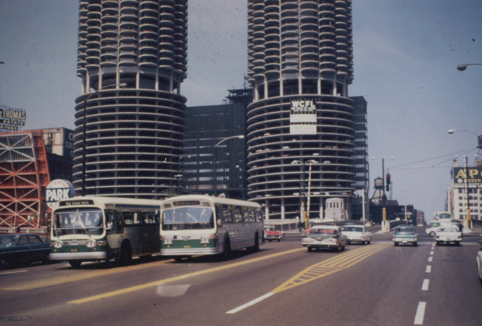 Two green and cream buses from the Chicago Transit Authority are on a multi-lane road, with the distinctive circular Marina City towers visible behind them under a clear sky.