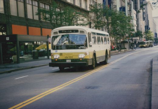 A white and yellow CTA bus with the number 130 on its front bumper drives down a city street, with pedestrians and another bus visible in the background.