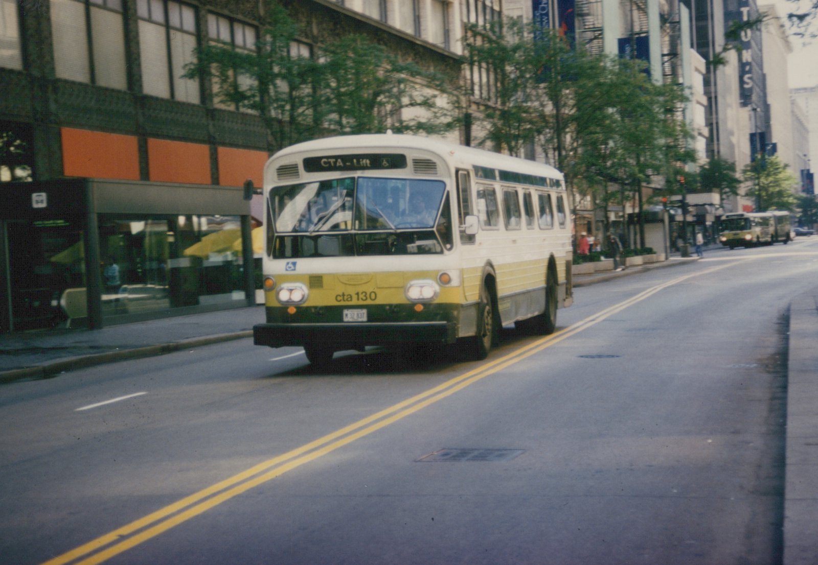 A white and yellow CTA bus with the number 130 on its front bumper drives down a city street, with pedestrians and another bus visible in the background.
