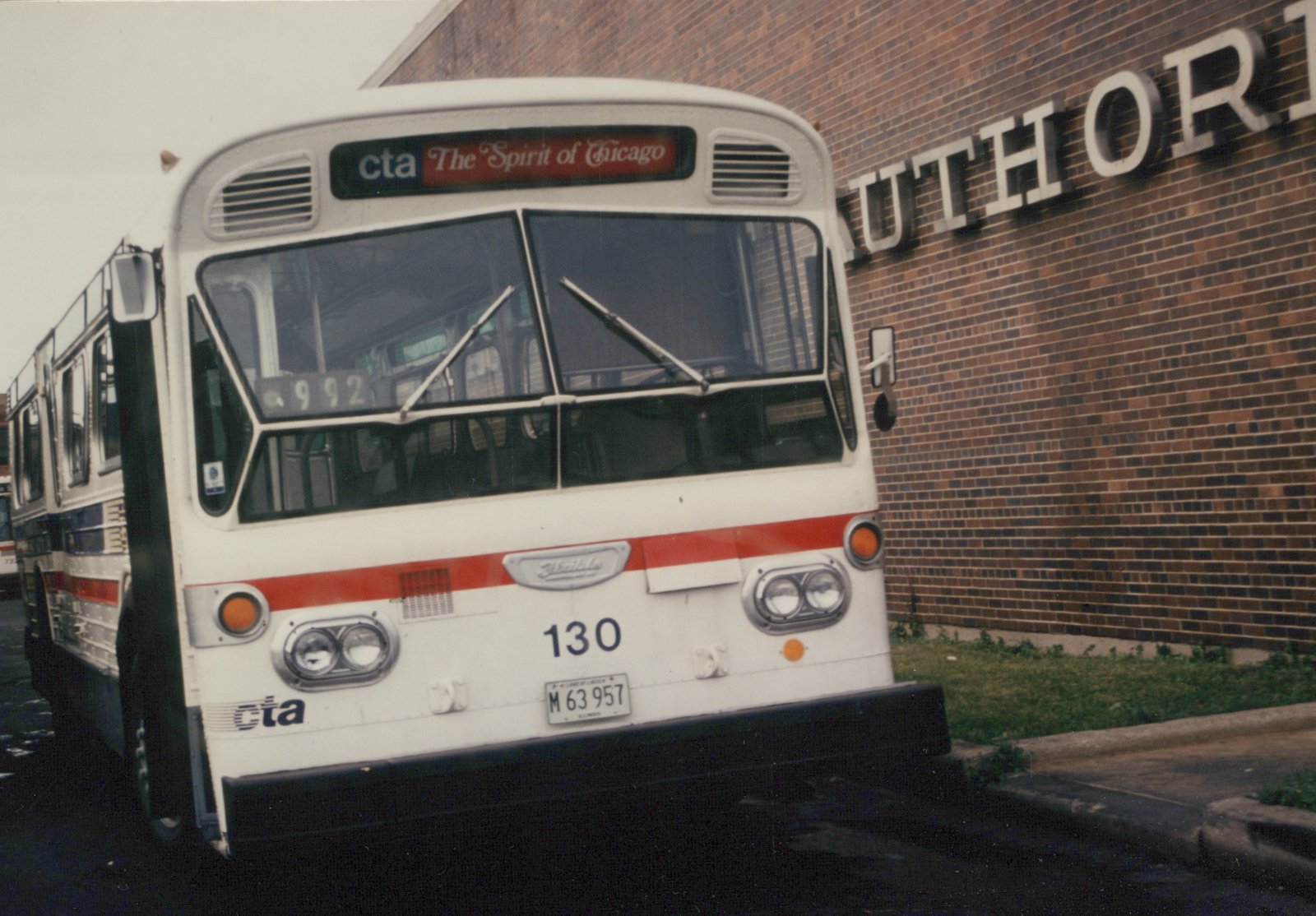 Front three-quarter view of a white and red CTA bus numbered 130, parked next to a brick building with the letters 'UTHOR' visible on it.