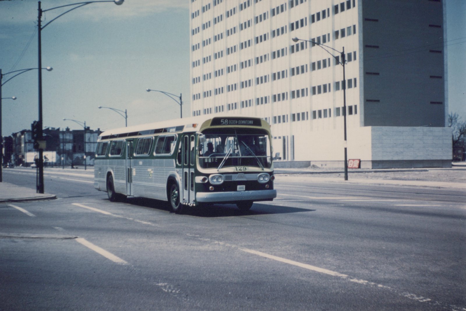 A green and cream CTA bus with the number 129 and route 58 Ogden-Downtown on its front sign, driving down a wide street with a large building in the background.