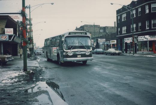 A vintage green and white bus with 'FREE SANTA BUS' on its sign drives down a snow-dusted city street lined with shops and cars from the 1970s.