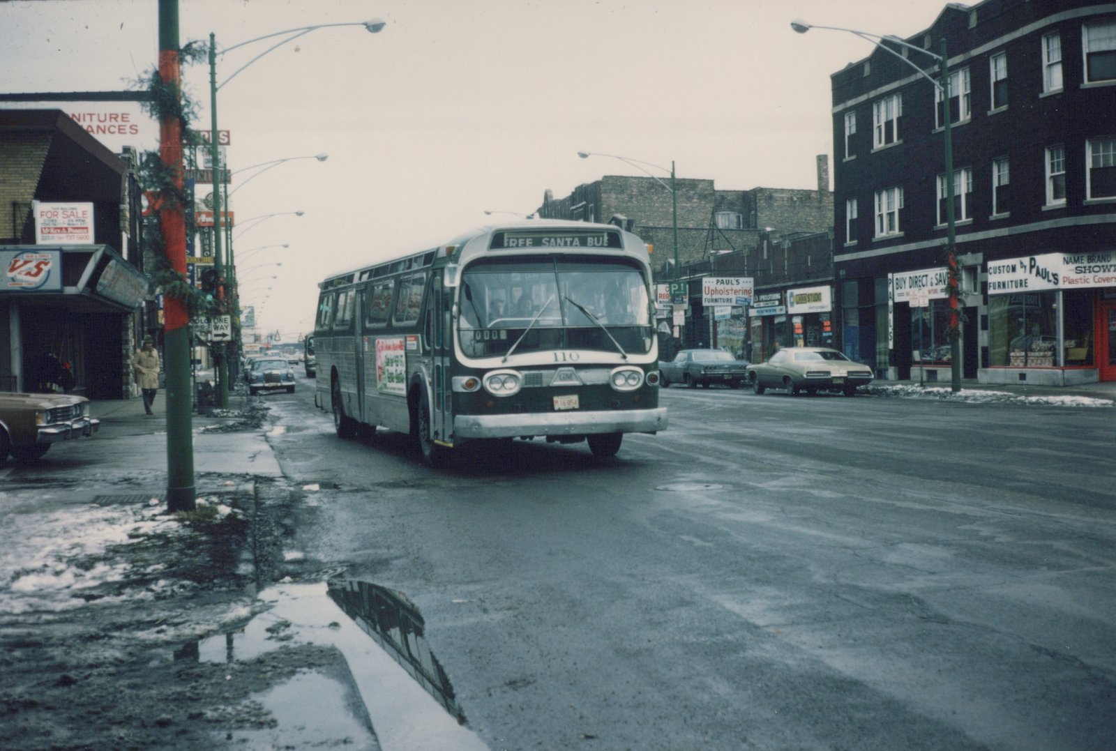A vintage green and white bus with 'FREE SANTA BUS' on its sign drives down a snow-dusted city street lined with shops and cars from the 1970s.