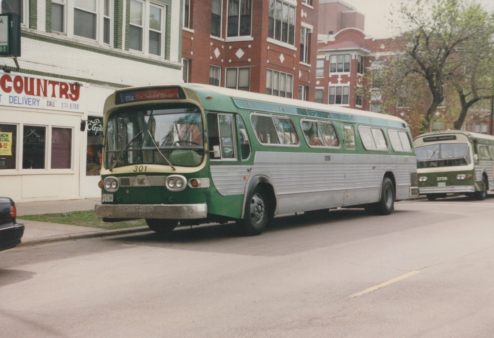 A green and silver Chicago Transit Authority bus, number 301, is parked on a city street with a second bus, number 3706, behind it. Buildings and a storefront are visible in the background.
