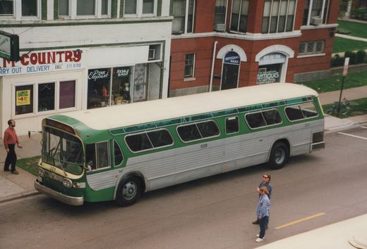 A green and silver CTA bus, number 301, is parked on a city street with three people nearby and shops in the background.