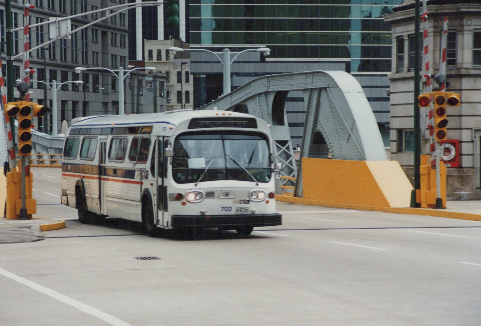 A white CTA bus numbered 702, with a blue and orange stripe, drives on a city street near a steel arch bridge, with traffic signals and modern buildings in the background.