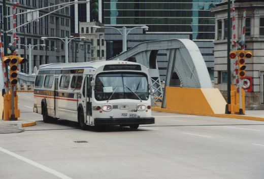 A white CTA bus numbered 702, with a blue and orange stripe, drives on a city street near a steel arch bridge, with traffic signals and modern buildings in the background.