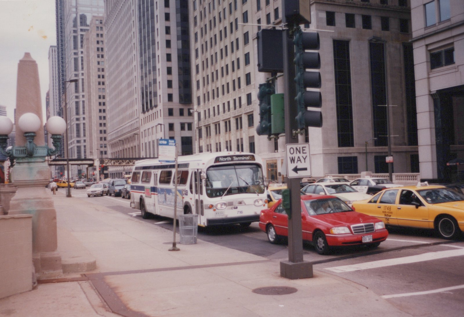 A white CTA bus with the number 716 and destination 'North Terminal' is stopped at a city intersection with tall buildings in the background; a red sedan and yellow taxi cab are in front of it, and a 'ONE WAY' street sign is visible.
