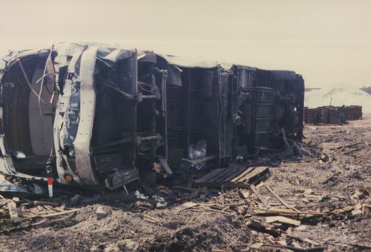 A destroyed and overturned bus or trolleybus in a yard, surrounded by rubble and barrels under an overcast sky.