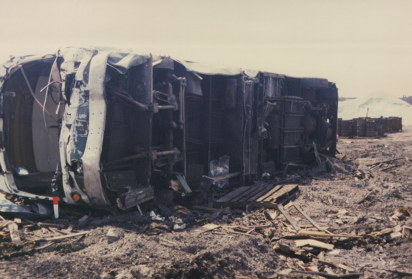 A destroyed and overturned bus or trolleybus in a yard, surrounded by rubble and barrels under an overcast sky.