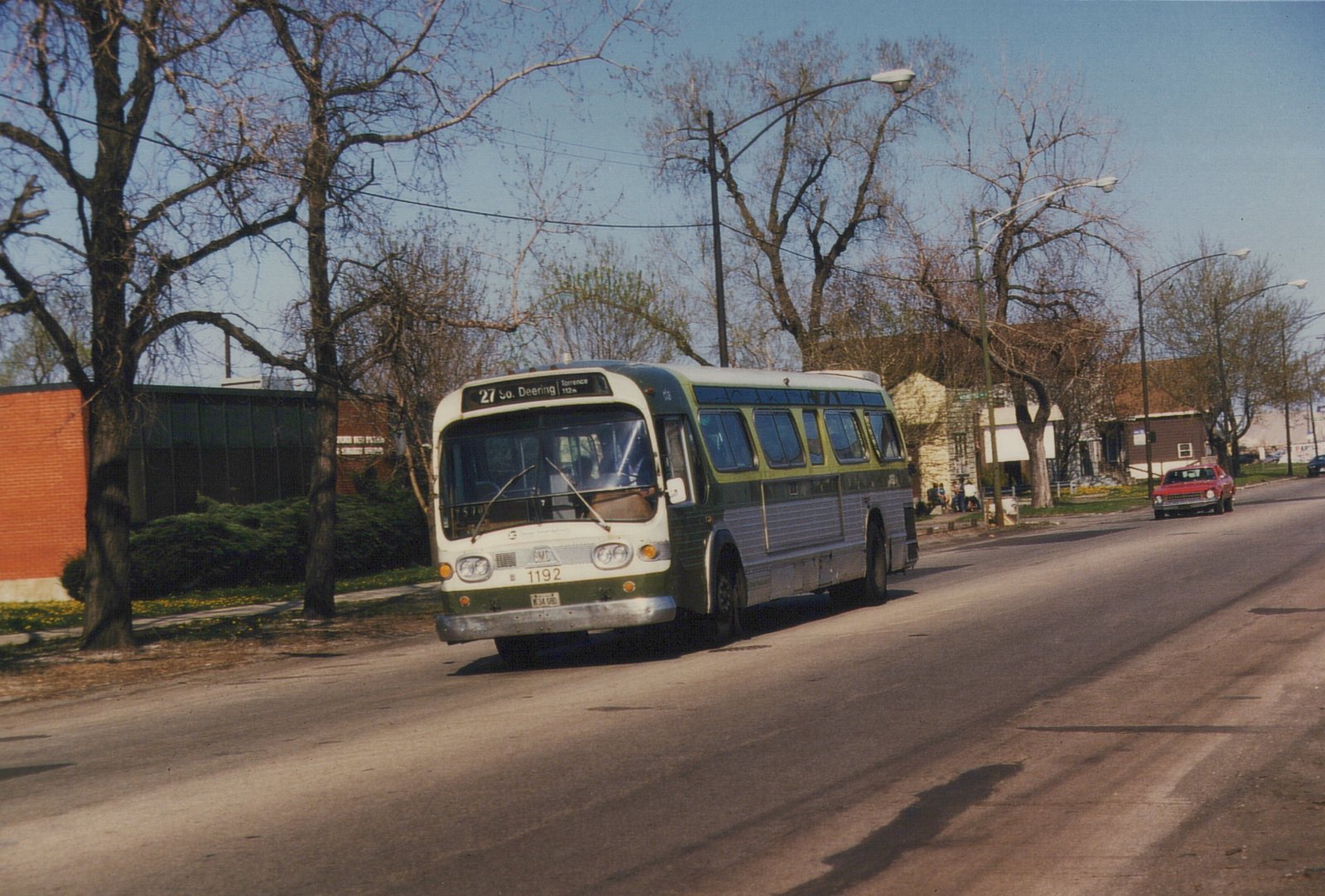 A green and cream CTA bus numbered 1192 drives down a sunny street, with bare trees and a red car in the background. The destination sign reads '27 So. Deering'.
