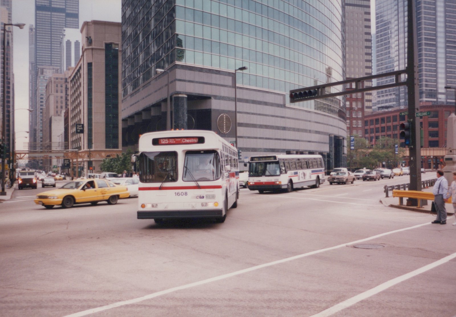 Two white CTA buses with red and blue stripes drive on a city street lined with tall buildings, with a yellow taxi cab nearby and pedestrians on the sidewalk.
