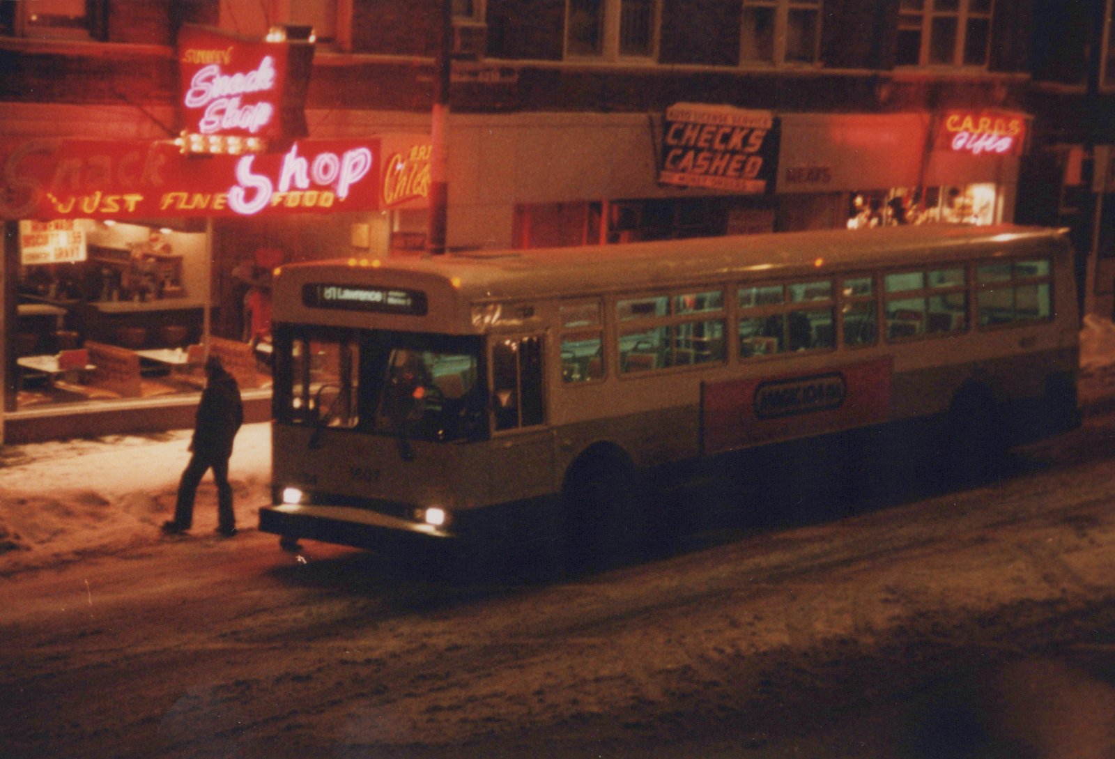 A night scene showing a CTA bus on a snowy street with a pedestrian walking past a brightly lit snack shop and check-cashing store.