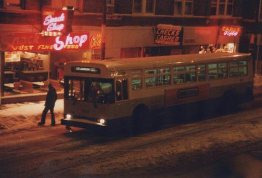A night scene showing a CTA bus on a snowy street with a pedestrian walking past a brightly lit snack shop and check-cashing store.
