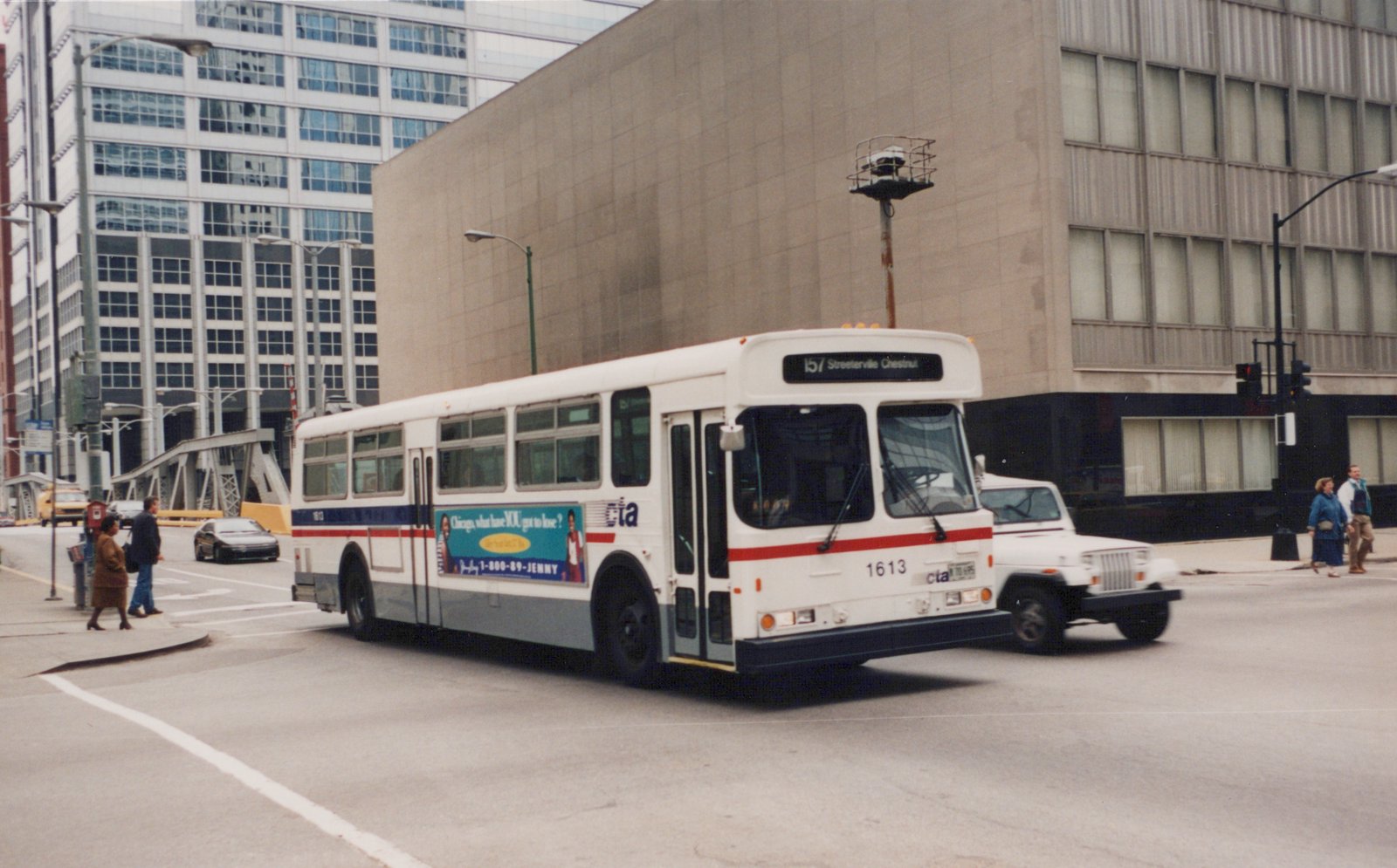 A white and gray Chicago Transit Authority bus numbered 1613 is turning at an intersection in downtown Chicago, with pedestrians and other vehicles nearby.