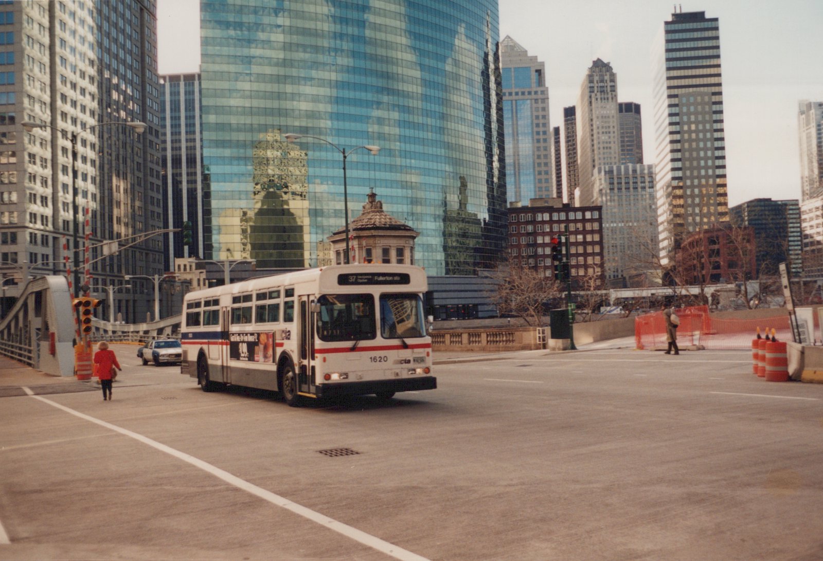 A white and red CTA bus numbered 1620 is turning on an urban street, with tall glass buildings in the background and a few pedestrians on the sidewalk.