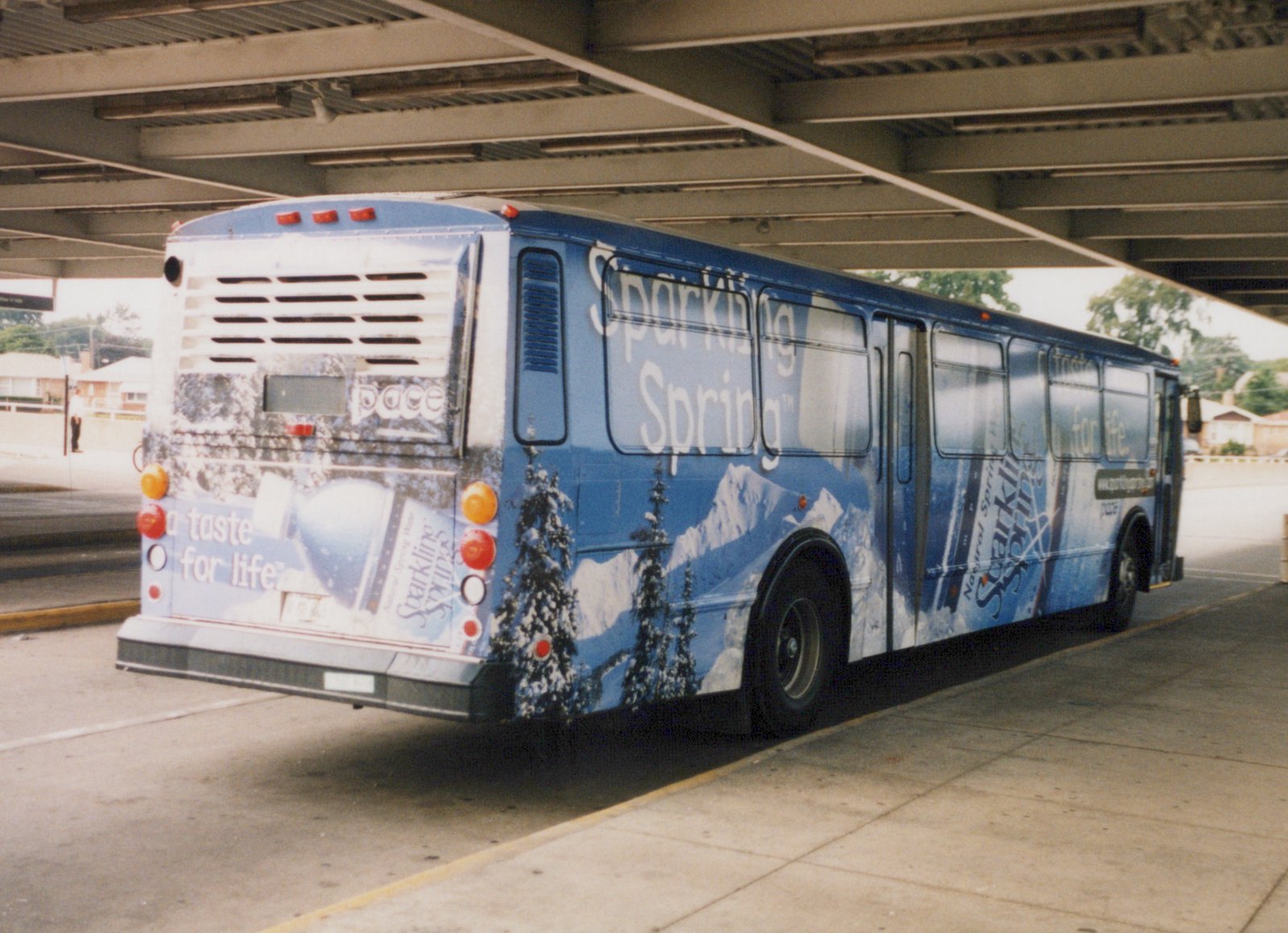 Rear three-quarter view of a blue Pace bus covered in a snow mountain advertisement for Sparkling Spring water, parked under a bridge on a sunny day.