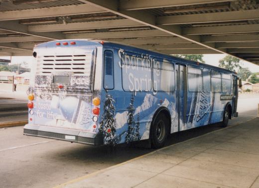 Rear three-quarter view of a blue Pace bus covered in a snow mountain advertisement for Sparkling Spring water, parked under a bridge on a sunny day.