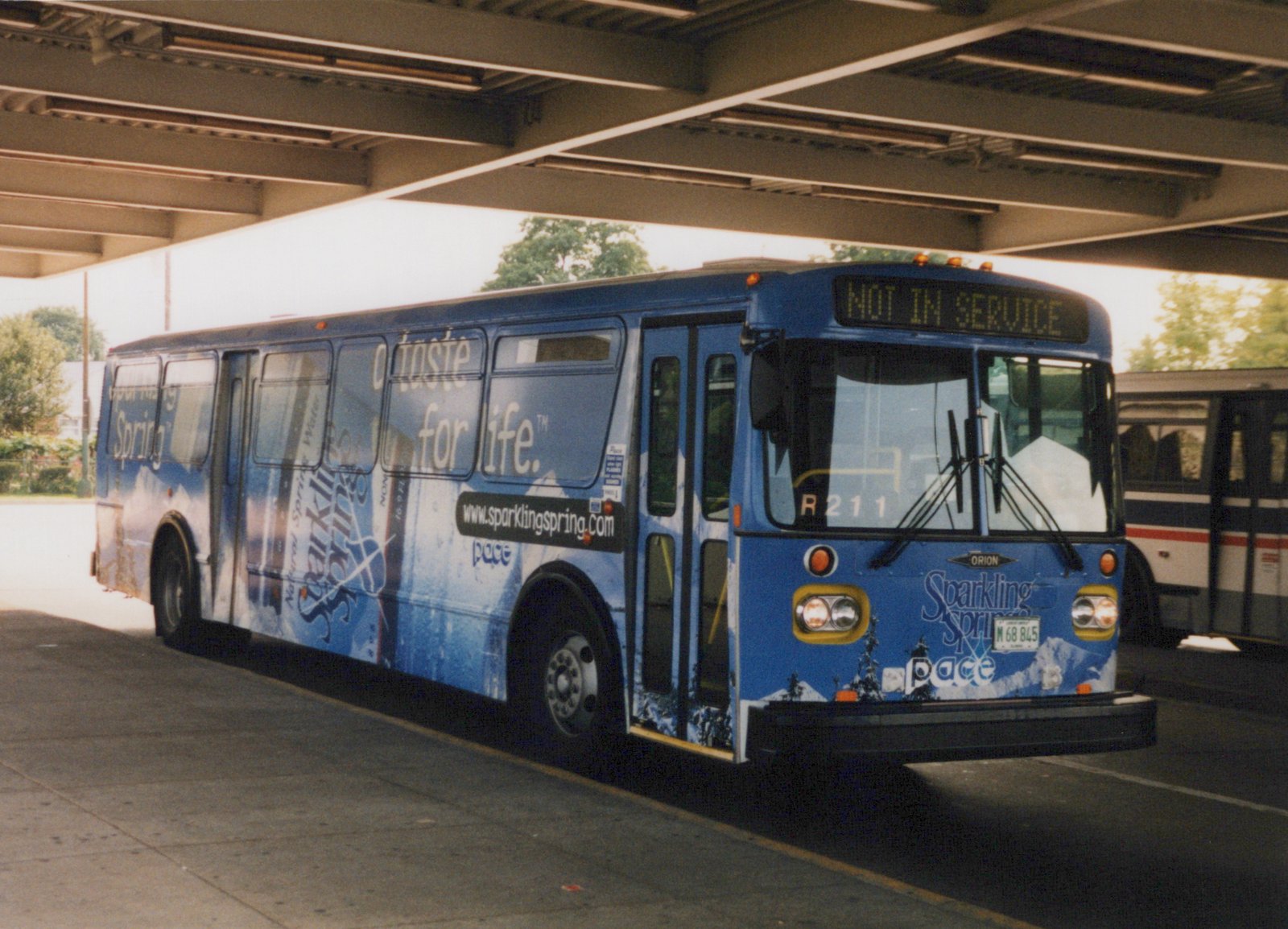 A blue Pace bus with a Sparkling Spring water advertisement, numbered R211, is parked under a concrete overpass with its destination sign reading 'NOT IN SERVICE'.