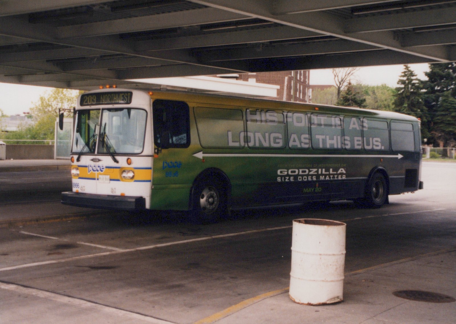 A green and white Pace bus with the number 8866 and a Godzilla movie ad is parked under a concrete overpass on a cloudy day.