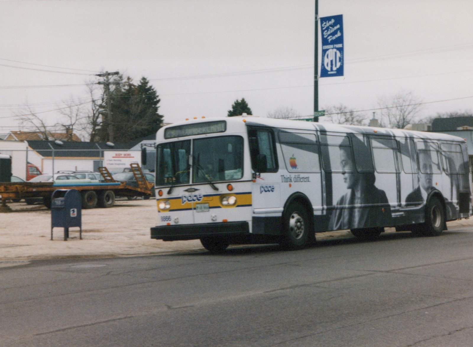 A white Pace bus with an Apple advertisement is driving down a street with a blue 'Shop Edison Park' banner on a pole nearby.