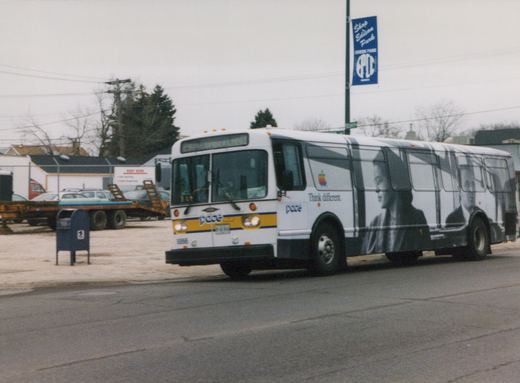 A white Pace bus with an Apple advertisement is driving down a street with a blue 'Shop Edison Park' banner on a pole nearby.