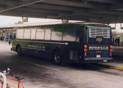 A dark green and black Pace bus is shown from the rear three-quarter view, parked under a structure with a Godzilla movie ad on its side and back; people are visible in the background.
