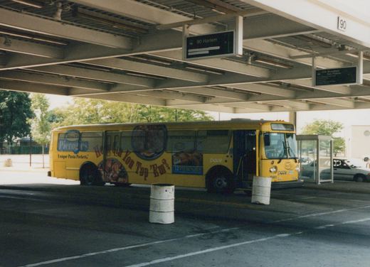 A yellow Pace bus with a large food ad is parked under the roof of a bus terminal, with overhead route signs for 90 Harlem and 209 Woodfield visible.