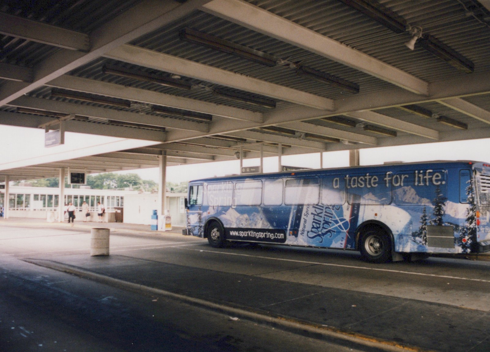 A blue bus with a Sparkling Spring water ad, parked under a covered terminal with a few people nearby.
