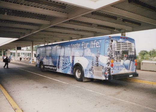 Side and rear view of a blue and white Pace bus wrapped in a Sparkling Spring ad, parked under a covered terminal structure with people walking nearby.