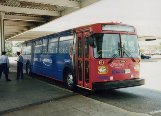 A red and blue Pace bus with the number 8869 is parked at a terminal under a shelter, with two people standing nearby.