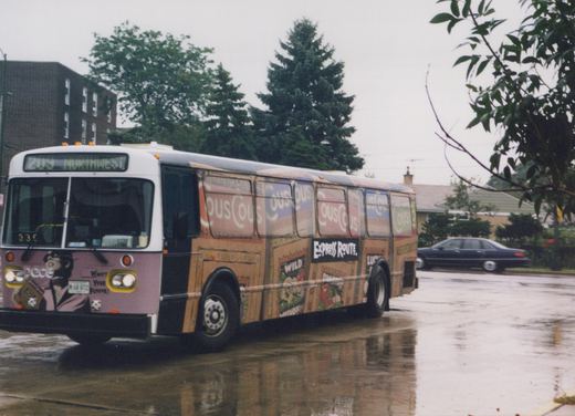 A purple and white Pace bus with a CousCous ad, number 330, displaying route 209 Northwest, parked on a wet street with trees and a building in the background.