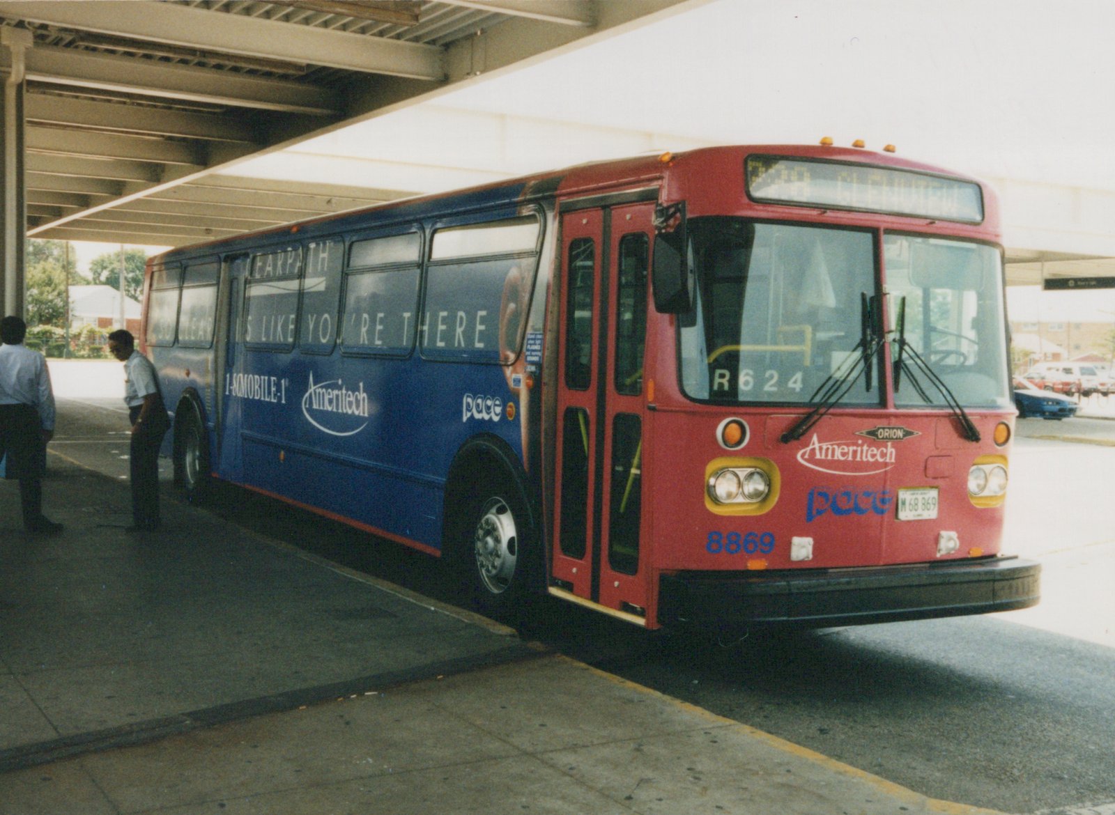 A red and blue Pace bus, number 8869, is parked under an overpass at a terminal with '228 GLENVIEW' on the front sign; two people are standing on the sidewalk nearby.