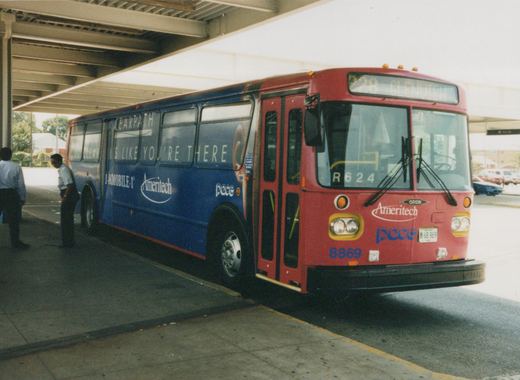 A red and blue Pace bus, number 8869, is parked under an overpass at a terminal with '228 GLENVIEW' on the front sign; two people are standing on the sidewalk nearby.