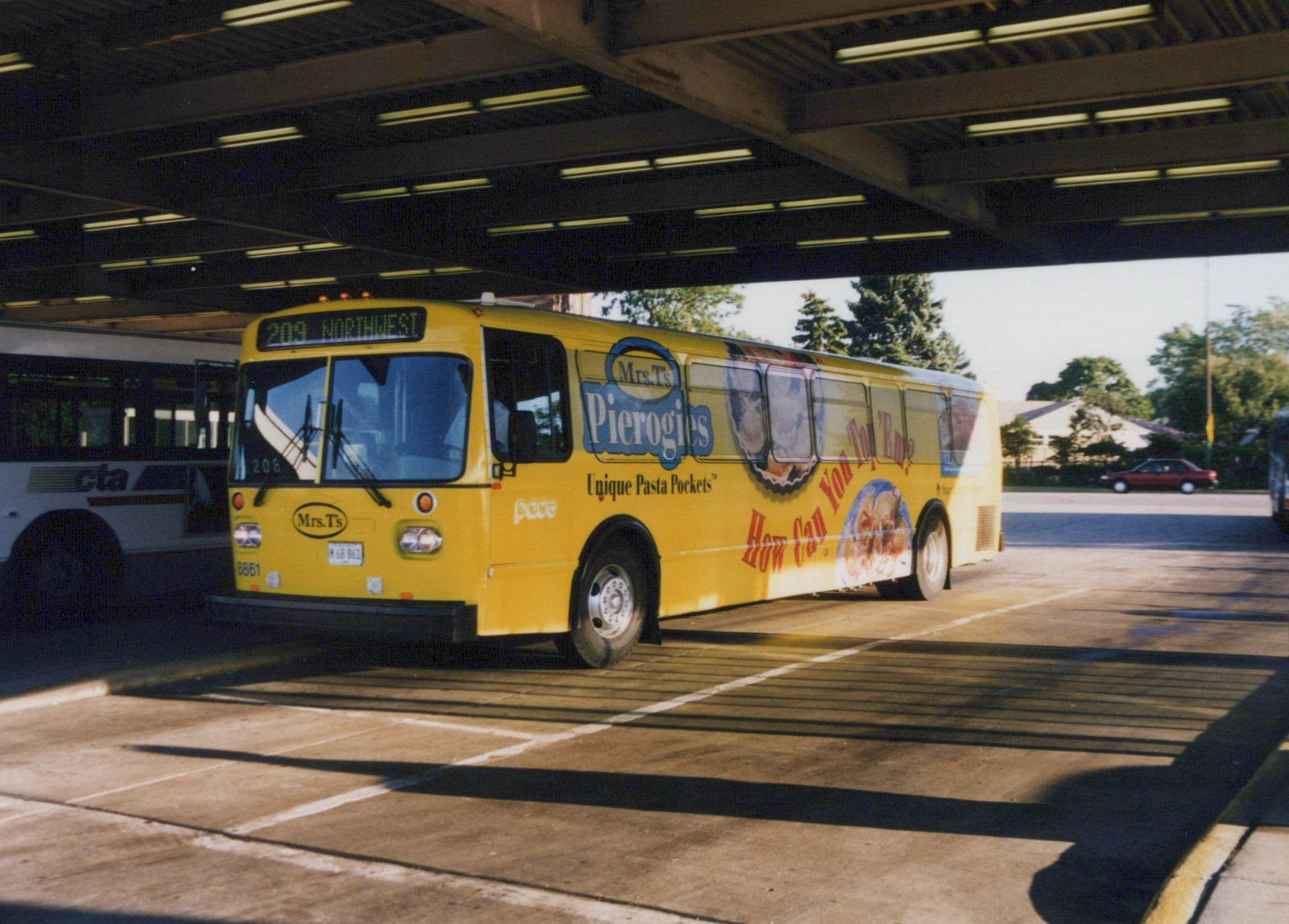 A yellow Pace bus, number 8861, with Mrs. T's Pierogies advertising on its side, is parked under a covered terminal roof, with a white CTA bus partially visible behind it.