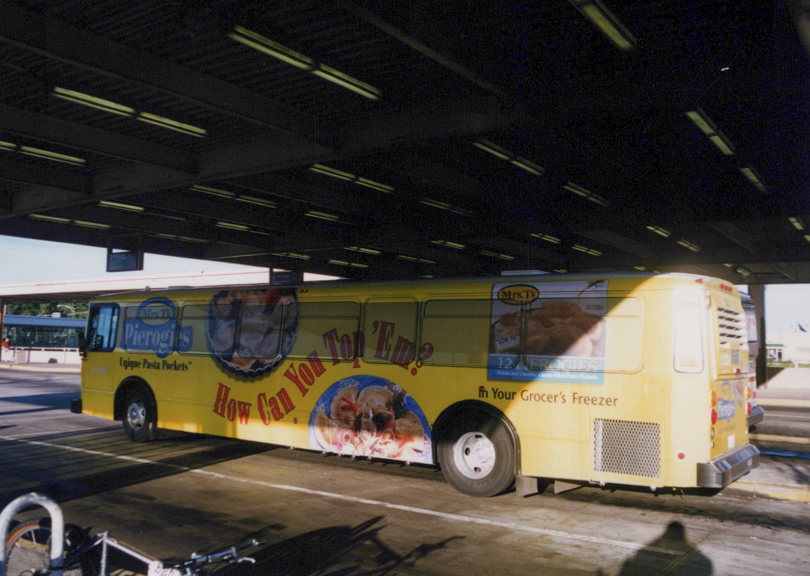 A yellow Pace bus with a Mrs. T's Pierogies ad parked under a shaded terminal canopy, viewed from a three-quarter rear angle.