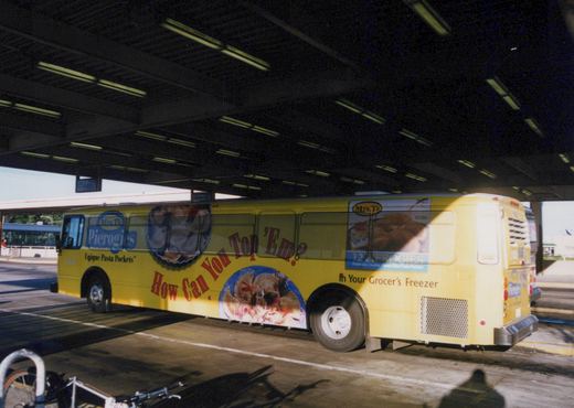 A yellow Pace bus with a Mrs. T's Pierogies ad parked under a shaded terminal canopy, viewed from a three-quarter rear angle.
