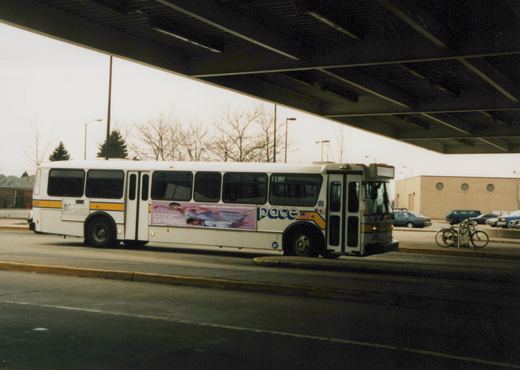 A white Pace bus with the number 2326 is parked under an overpass. The side of the bus features an advertisement for The Joffrey Ballet of Chicago. Several cars and a bicycle are visible in the background.