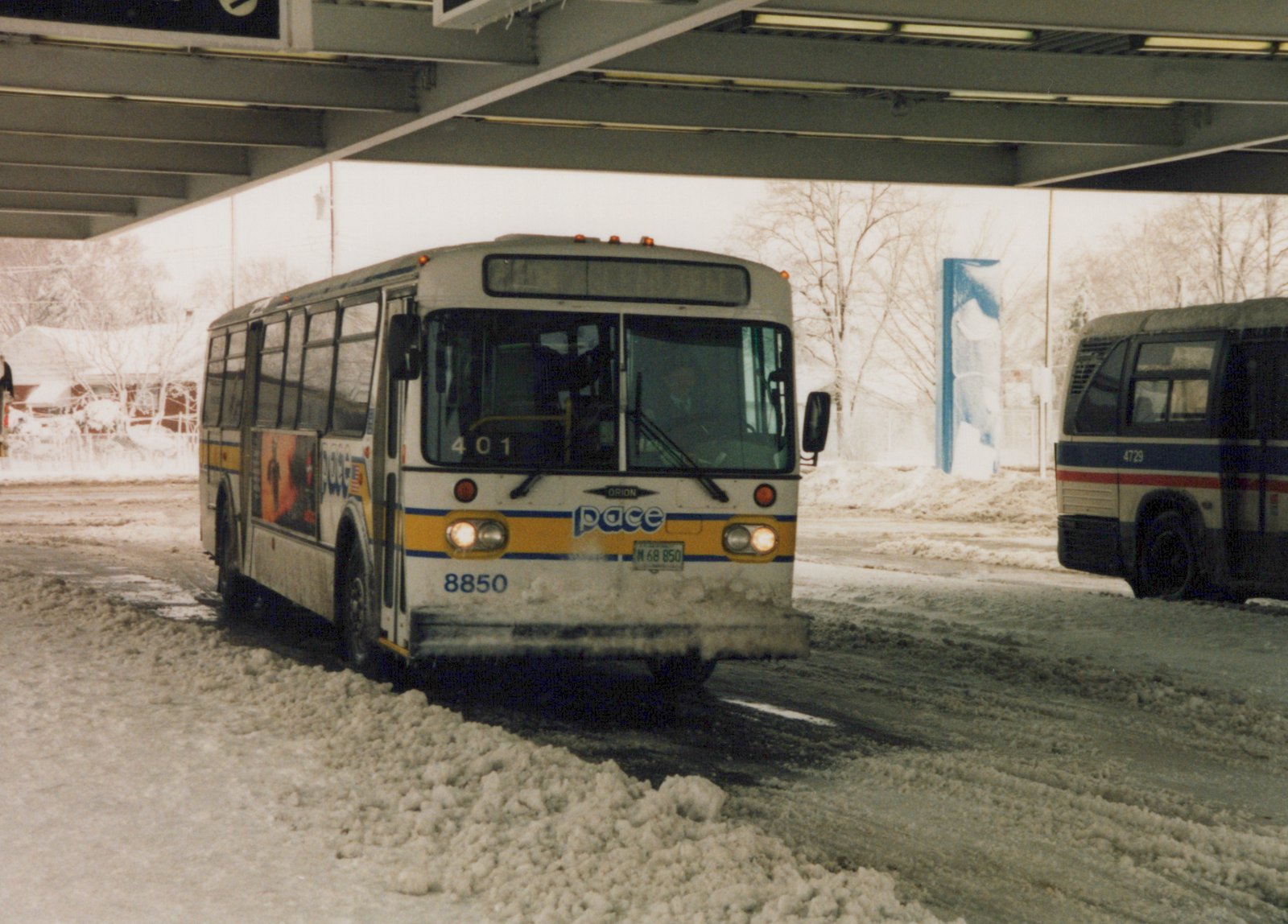 Two Pace buses, one with the number 8850 on its bumper and another with 4729 on its side, are on a snow-covered street under an overpass, with bare trees in the background.