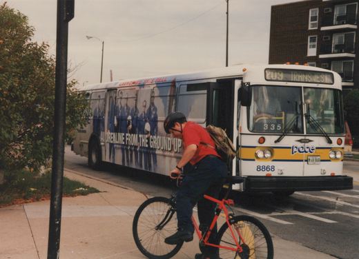 A white and yellow Pace bus with the number 8868 is stopped on a city street, with a cyclist on a red bicycle riding on the sidewalk in front of it.