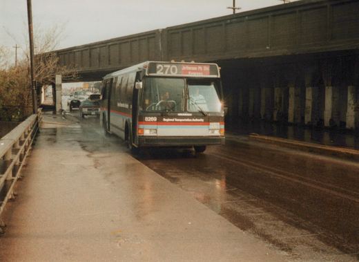A brown, white, red, and blueRegional Transportation Authority motor bus, number 8269, travels down a wet street under a dark overpass on a cloudy day.
