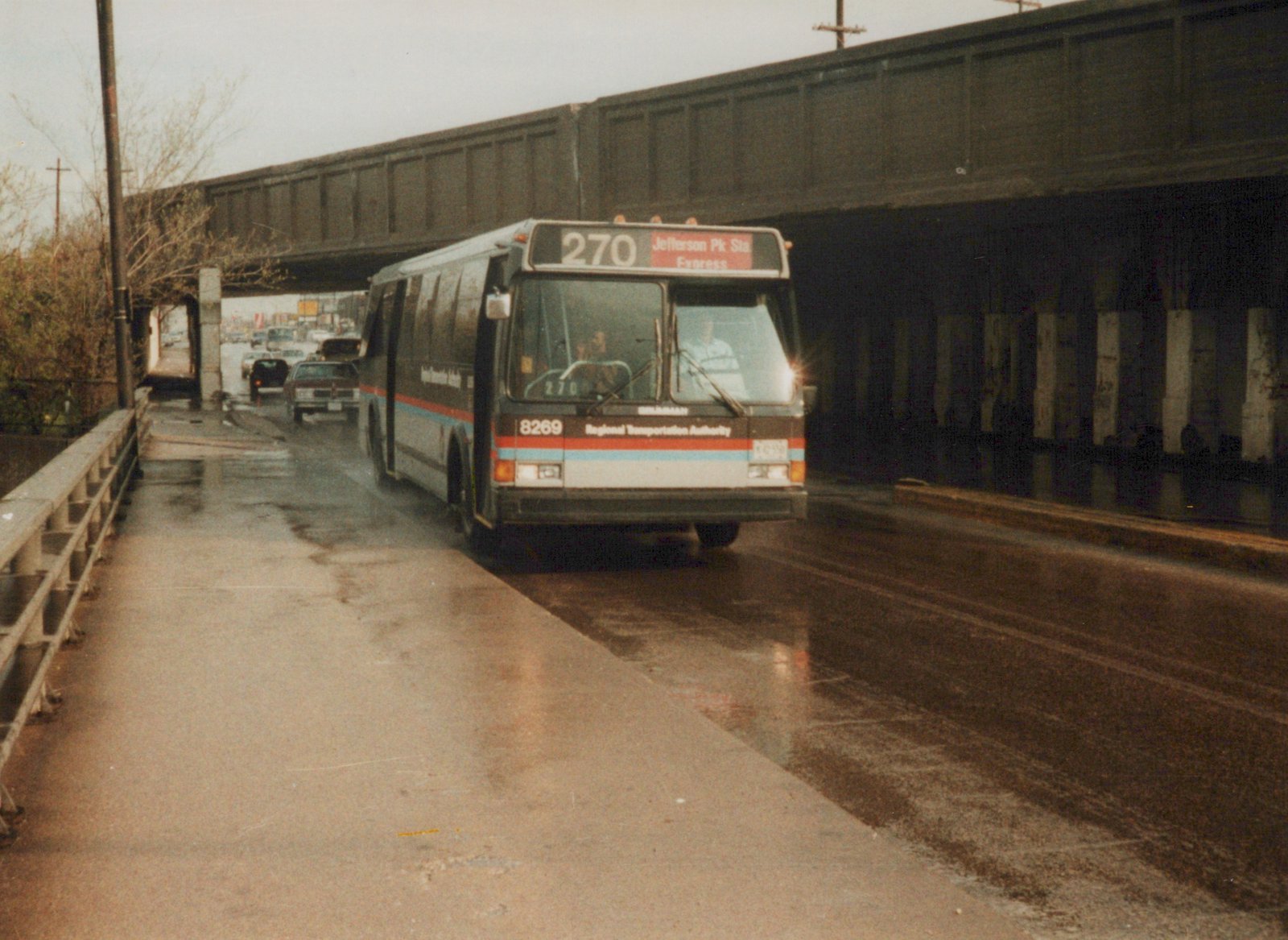 A brown, white, red, and blueRegional Transportation Authority motor bus, number 8269, travels down a wet street under a dark overpass on a cloudy day.