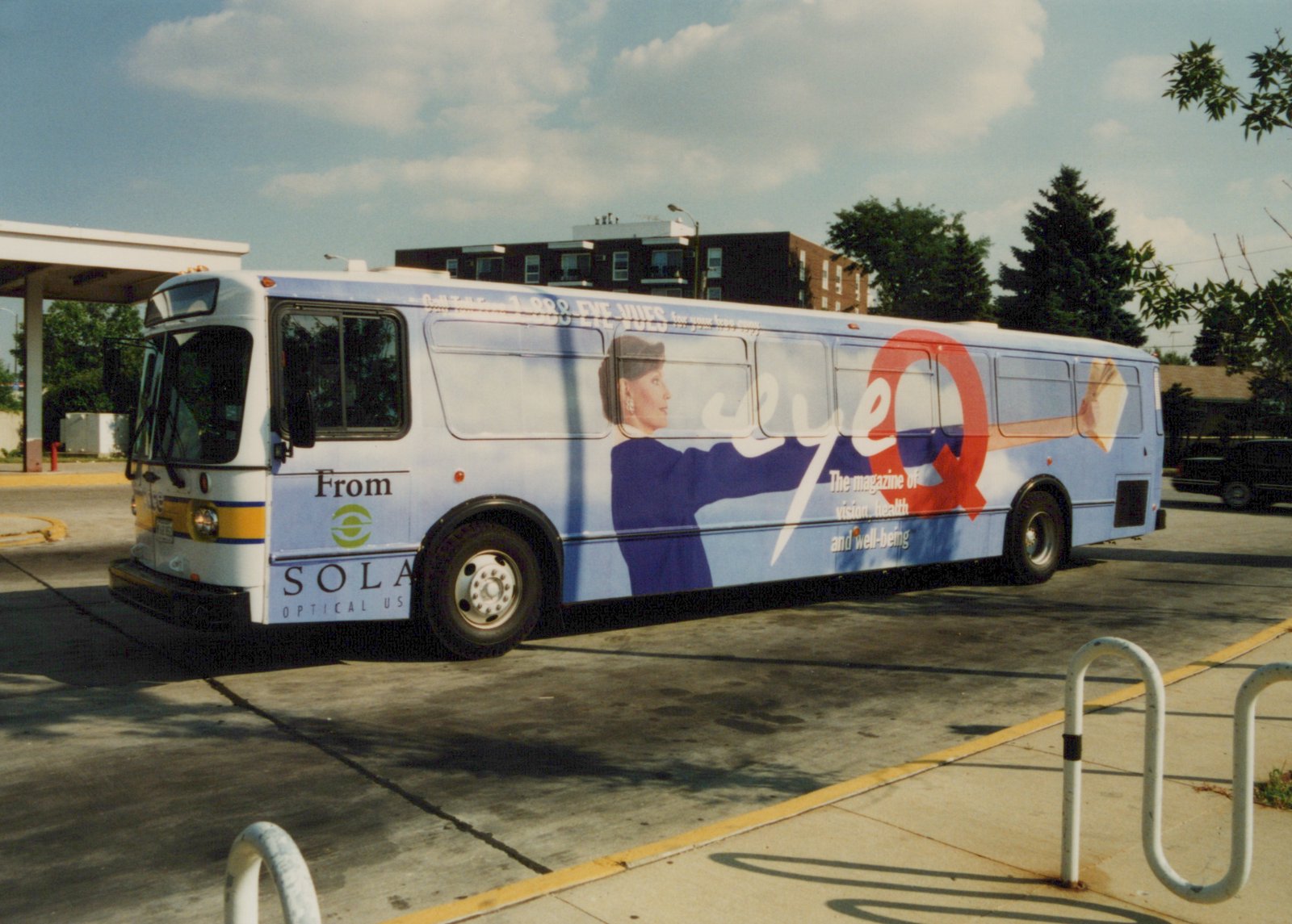 Side view of a white Pace bus with yellow and blue stripes, featuring a large 'Eye Q' ad, parked on a sunny day with buildings in the background.