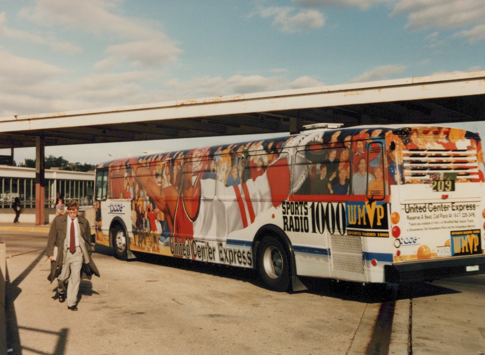 A white Pace bus with a colorful advertisement for the United Center Express and Sports Radio 1000 WMVP, number 209, is parked at a covered station platform on a sunny day.