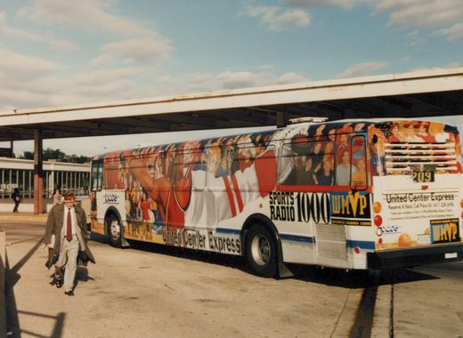 A white Pace bus with a colorful advertisement for the United Center Express and Sports Radio 1000 WMVP, number 209, is parked at a covered station platform on a sunny day.