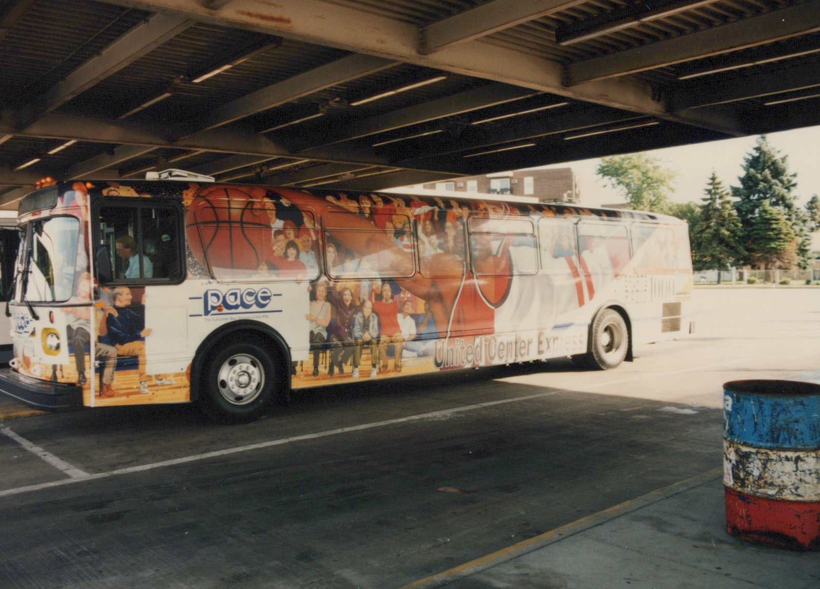 A white Pace Motor bus with a colorful basketball-themed wrap and 'United Center Express' lettering is parked under a bridge on a sunny day, with two people visible inside.