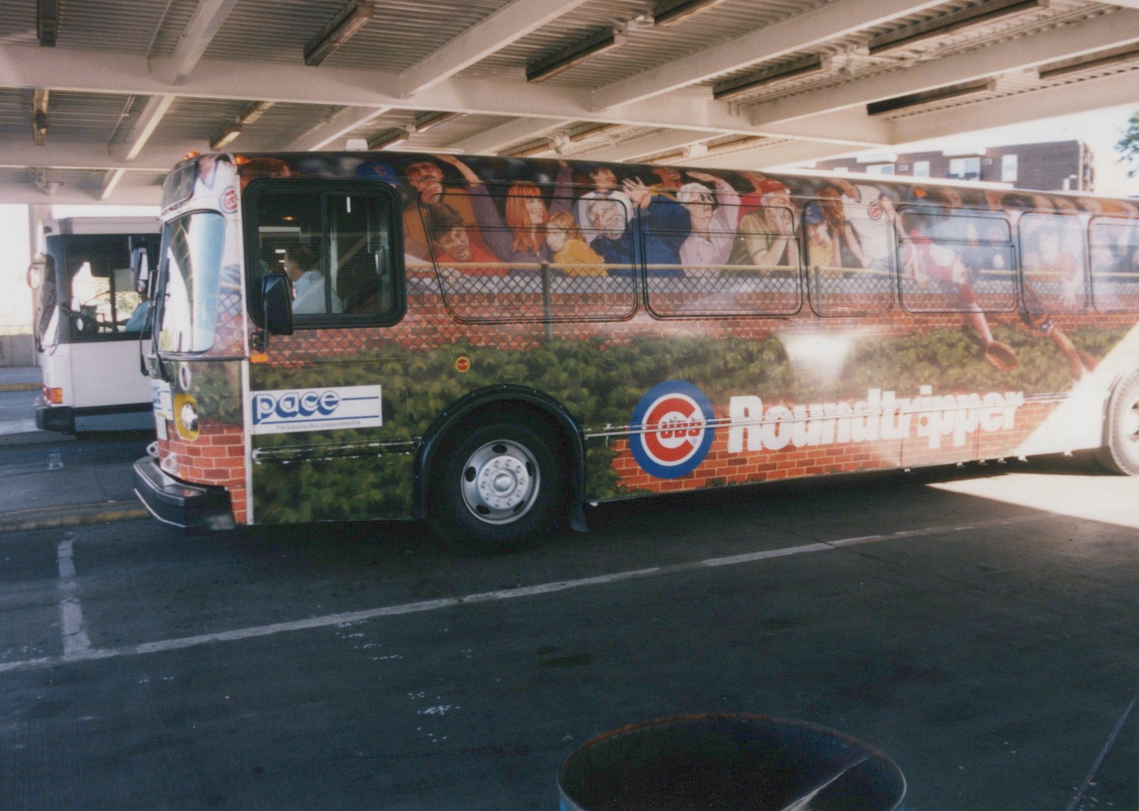 A Pace motor bus with a Chicago Cubs 'Roundtripper' advertisement wrapped around its side, parked on a paved area under a covered structure.