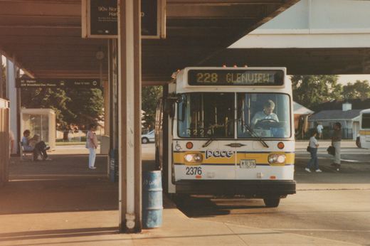 Front view of a white Pace bus with yellow and blue stripes, number 2376, stopped at a covered bus terminal with people waiting on the platform.